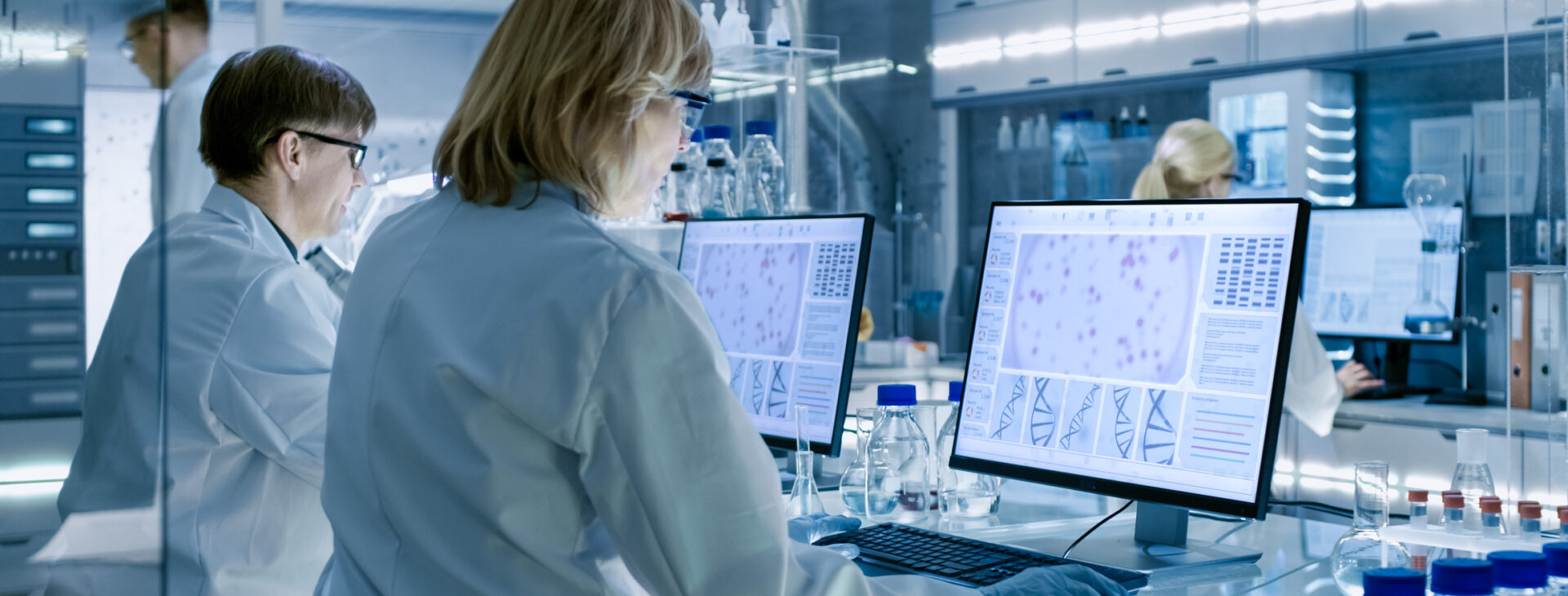 Female and Male Scientists Working on their Computers In Big Modern Laboratory. Various Shelves with Beakers, Chemicals and Different Technical Equipment is Visible.