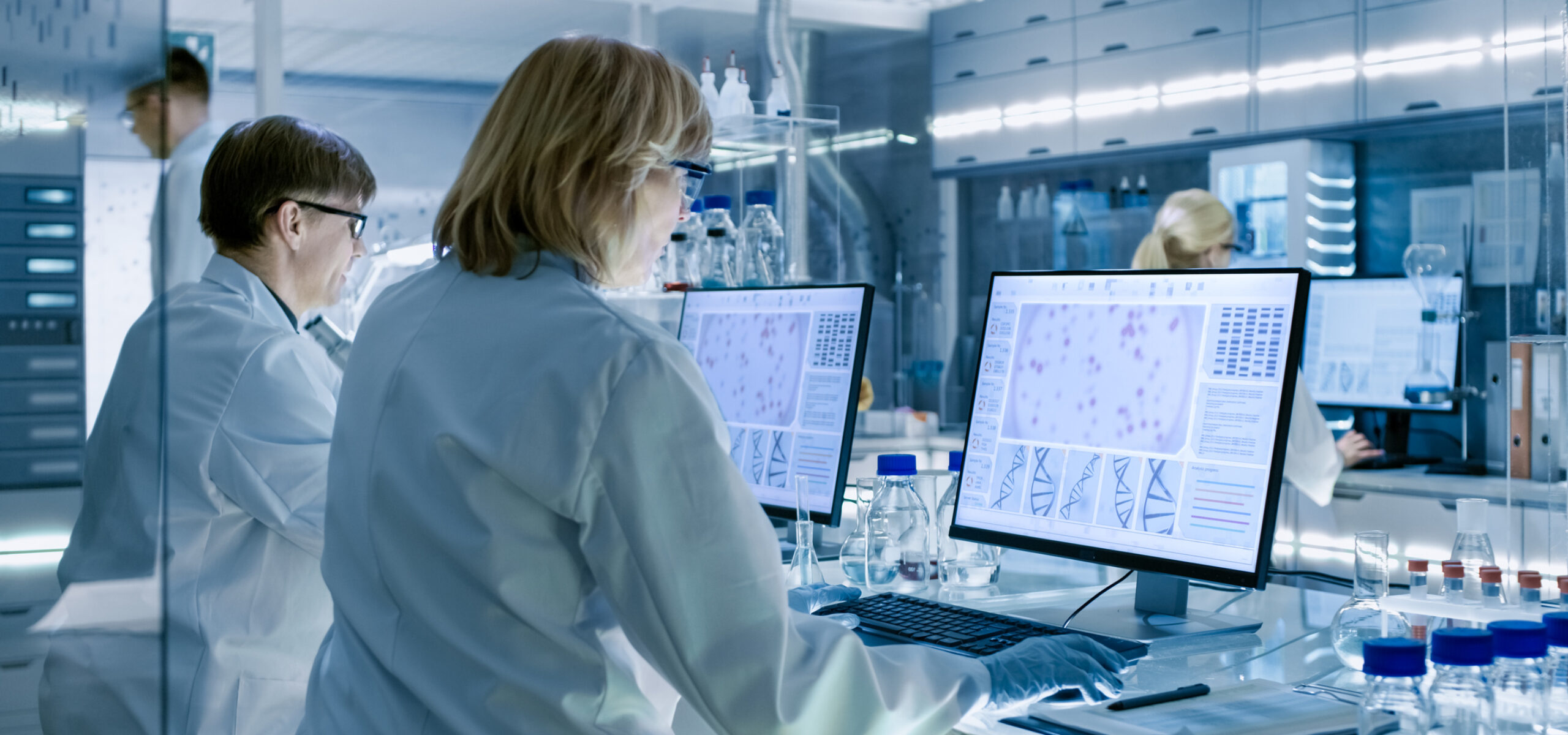 Female and Male Scientists Working on their Computers In Big Modern Laboratory. Various Shelves with Beakers, Chemicals and Different Technical Equipment is Visible.